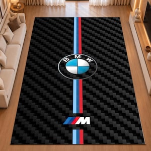 Op de afbeelding: Een rechthoekig vloerkleed met een zwarte koolstofvezelpatroon achtergrond en een verticale lichtblauwe, donkerblauwe en rode streep door het midden. Het BMW-logo en het M-embleem zijn in het midden van het vloerkleedontwerp gedrukt.