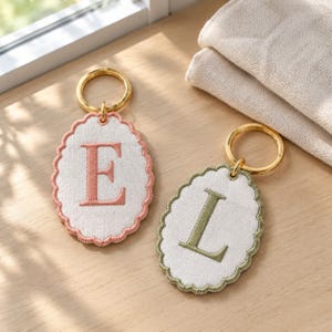 May include: Two oval-shaped fabric keychains with gold-toned metal rings. One keychain features a coral-coloured embroidered letter E with a matching scalloped edge, while the other displays an olive green embroidered letter L with a matching scalloped edge.