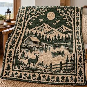 May include: A rectangular crochet blanket featuring a dark green and cream landscape scene. The design depicts a cabin, mountains, a lake with a canoe, and a deer. The border is decorated with a repeating pattern of small pine trees and leaves.