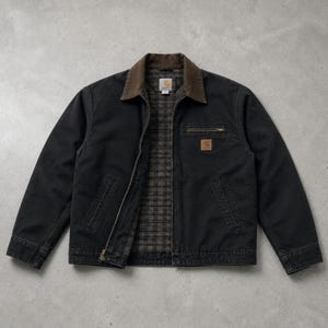 May include: A black canvas jacket with a brown corduroy collar and a plaid interior lining. The jacket features a front zip fastening, two lower welt pockets, and a zipped chest pocket with a square Carhartt brand patch on the left side.