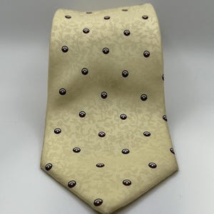 May include: A pale yellow necktie featuring a subtle, tonal floral jacquard pattern throughout the fabric. The tie is decorated with a repeating pattern of small, dark red and white circular icons arranged in a grid across the entire surface.