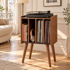 May include: A wooden record player stand with four vertical storage compartments. The top surface holds a black turntable. The compartments contain vinyl records and a pair of tan and silver headphones. The unit features four tapered legs.