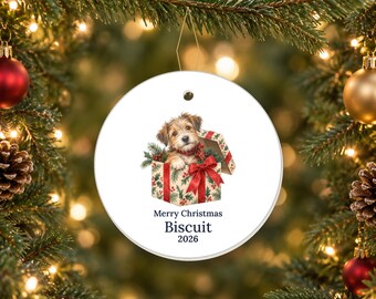 May include: A round white ceramic Christmas ornament featuring a brown and white terrier puppy sitting inside a gift box with a red bow. The text reads Merry Christmas, Biscuit, and 2026 in dark blue lettering centered below the illustration.