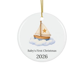 May include: A round white ceramic ornament featuring a gold hanging string. The center displays a wooden sailboat with a gold star on its sail, resting on a blue-tinted cloud. Below the illustration, the text reads Baby's First Christmas 2026.