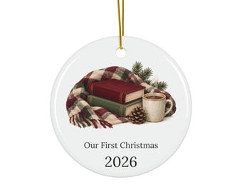 May include: A round white ceramic ornament hanging from a gold string. The design features a plaid scarf, a red book, a green book, a pinecone, a pine branch, and a mug of hot beverage. The text reads Our First Christmas and the year 2026.