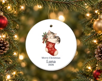 May include: A round white ceramic Christmas ornament featuring a sleeping tabby kitten inside a red stocking with white snowflake patterns and a sprig of holly. The text reads Merry Christmas, Luna, and 2026 in dark blue lettering.