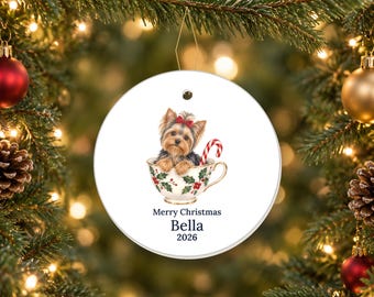 May include: A round white ceramic Christmas ornament featuring a Yorkshire Terrier puppy with a red bow sitting in a teacup decorated with holly leaves and berries, next to a candy cane. Text reads Merry Christmas, Bella, 2026.