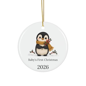 Puede incluir: Un adorno redondo de cerámica blanca con un pingüino blanco y negro que lleva una bufanda color mostaza y sostiene una rama de bayas rojas. El texto Baby's First Christmas y el año 2026 están impresos en negro debajo del pingüino.