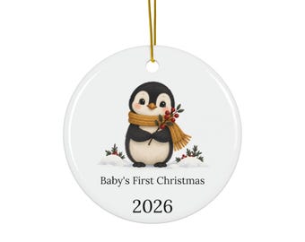 May include: A round white ceramic ornament featuring a black and white penguin wearing a mustard yellow scarf and holding a sprig of red berries. The text Baby's First Christmas and the year 2026 are printed in black below the penguin illustration.