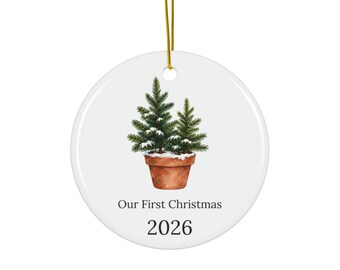 May include: A round white ceramic ornament featuring a watercolor illustration of two small evergreen trees in a terracotta pot with snow on the branches. The text reads Our First Christmas and the year 2026 in black font. Includes a gold string.