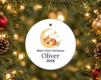 May include: A round white ceramic ornament featuring a watercolor illustration of a fox sleeping on a crescent moon with stars. The text reads Baby's First Christmas, the name Oliver, and the year 2026 in dark blue lettering on the front surface.