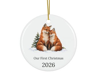 May include: A round white ceramic ornament featuring a detailed illustration of two foxes sitting together in the snow near a small pine tree. The text "Our First Christmas" and the year "2026" are printed in black below the foxes. A gold string is attached.