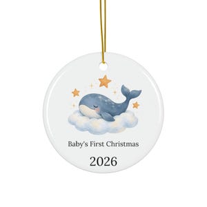 Puede incluir: Un adorno redondo de cerámica blanca que cuelga de un cordón dorado. El centro muestra una ballena azul sobre una nube azul claro rodeada de estrellas naranjas. Debajo de la ilustración, el texto dice Baby's First Christmas y el año 2026.