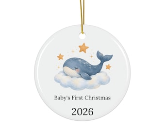 May include: A round white ceramic ornament hanging from a gold string. The center features a blue whale resting on a light blue cloud surrounded by orange stars. Below the illustration, the text reads Baby's First Christmas and the year 2026.