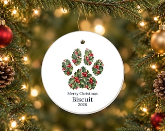 May include: A round white ceramic ornament featuring a paw print design composed of green holly leaves and red berries. Below the paw print, the text reads Merry Christmas, Biscuit, and the year 2026 in a dark blue serif font.