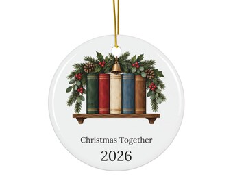 May include: A round white ceramic ornament featuring a shelf with five colorful books, pine branches, pinecones, red berries, and a small gold bell. Below the shelf, the text reads Christmas Together and the year 2026 in a black serif font.
