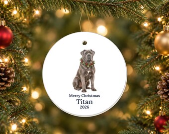 May include: A round white ceramic ornament featuring a gray Cane Corso dog wearing a green and red holly wreath around its neck. Below the dog, the text reads Merry Christmas, Titan, and 2026 in a dark blue serif font.