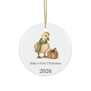 Puede incluir: Un adorno redondo de cerámica blanca colgado de un cordón dorado. Muestra un patito amarillo esponjoso con una bufanda verde junto a una caja de regalo marrón con un lazo rojo. El texto dice Baby's First Christmas y el año 2026.