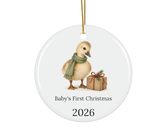 May include: A round white ceramic ornament hanging from a gold string. It features a fluffy yellow duckling wearing a green scarf next to a brown gift box with a red ribbon. The text reads Baby's First Christmas and the year 2026.