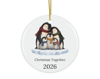 May include: A round white ceramic ornament featuring an illustration of four penguins huddled together on an ice floe, wearing colorful scarves. The text reads Christmas Together and 2026. A gold string is attached to the top for hanging.