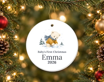 May include: A round white ceramic ornament featuring a watercolour illustration of a polar bear cub wearing a yellow jumper and blue scarf, playing in the snow. The text reads Baby's First Christmas, the name Emma, and the year 2026 in dark blue.