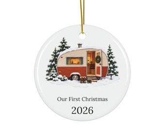 May include: A round white ceramic ornament featuring a vintage-style camper trailer in a snowy forest setting. The camper is red and cream with an open door and a wreath. The text reads Our First Christmas and the year 2026 in black font.