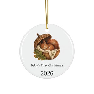 Puede incluir: Un adorno redondo de cerámica blanca colgado de un cordón dorado. En el centro, una ardilla marrón duerme dentro de una gran bellota forrada con una manta color crema, junto a una hoja de roble. El texto dice Baby's First Christmas y 2026.