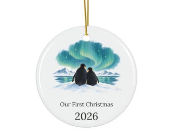 May include: A round white ceramic ornament featuring a gold hanging string. The illustration depicts two penguins sitting on snow, looking at a green aurora borealis over mountains and water. The text reads Our First Christmas and the year 2026.