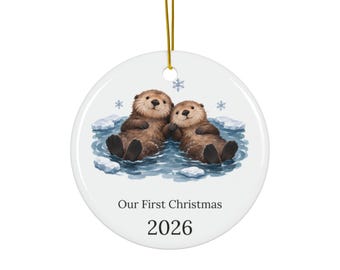 May include: A round white ceramic ornament featuring a watercolor illustration of two brown sea otters floating in blue water with ice chunks and snowflakes. The text "Our First Christmas" and "2026" is printed in black below the otters.