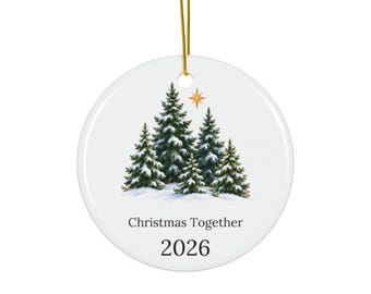 May include: A round white ceramic ornament hanging from a gold string. The center features a printed illustration of four snow-covered evergreen trees under a gold star. Below the trees, the text reads Christmas Together and the year 2026.