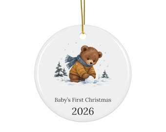 May include: A round white ceramic ornament hanging from a gold string. The center features a brown bear wearing a yellow sweater and blue scarf, playing in the snow near small pine trees. Below the illustration, the text reads Baby's First Christmas 2026.