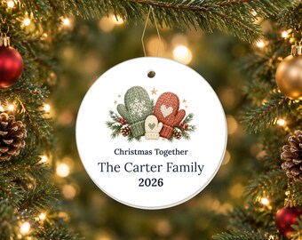 May include: A round white ceramic Christmas ornament featuring a graphic of three knitted mittens in green, red, and cream colors, surrounded by pine branches and berries. The text reads Christmas Together, The Carter Family, and 2026.