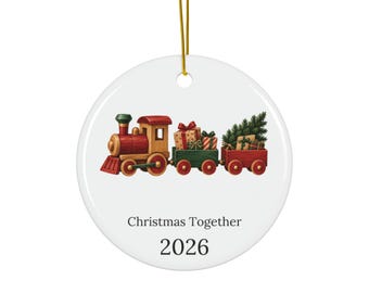 May include: A round white ceramic ornament featuring a red and green toy train carrying wrapped gifts and a small pine tree. The text "Christmas Together" is printed in black above the year "2026". A gold string is attached to the top.