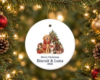 May include: A round white ceramic Christmas ornament featuring a golden retriever wearing a red scarf and a tabby cat with a red bow sitting by a decorated tree and gifts. The text reads Merry Christmas, Biscuit & Luna, 2026.