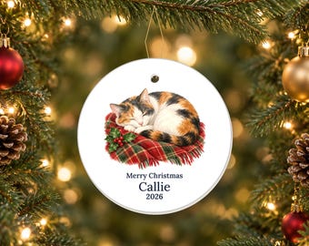 May include: A round white ceramic Christmas ornament featuring a sleeping calico cat curled up on a red and green plaid blanket with holly berries. The text Merry Christmas, Callie, and 2026 is printed in dark blue below the illustration.