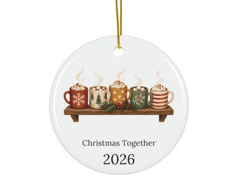 May include: A round white ceramic ornament hanging from a gold string. The design features five mugs of hot cocoa on a wooden shelf, with the text "Christmas Together" and "2026" printed below the illustration in a dark grey font.