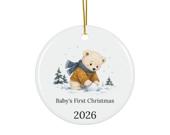 May include: A round white ceramic ornament hanging from a gold string. The center features a watercolor illustration of a light brown bear wearing a blue scarf and orange sweater, playing in the snow. Text reads Baby's First Christmas and 2026.