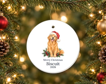 May include: A round white ceramic ornament featuring a golden retriever wearing a red Santa hat, sitting amidst holly leaves and red berries. Below the dog, the text reads Merry Christmas, Biscuit, and the year 2026 in dark blue font.