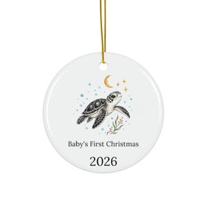 Puede incluir: Un adorno redondo de cerámica blanca colgado de un cordón dorado. El centro presenta una ilustración de una tortuga marina en blanco y negro con burbujas azules, una luna creciente dorada, estrellas y vegetación. El texto dice Baby's First Christmas y 2026.