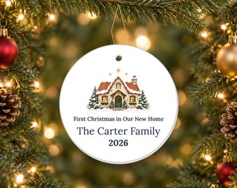 May include: A round white ceramic ornament featuring a colorful illustration of a snow-covered cottage with a red roof and pine trees. The text reads First Christmas in Our New Home, The Carter Family, and 2026 in dark blue lettering.