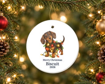 May include: A round white ceramic ornament featuring a brown dachshund wrapped in colorful Christmas lights. Below the dog, the text reads Merry Christmas, Biscuit, and 2026. The ornament hangs from a gold string against a blurred evergreen tree.
