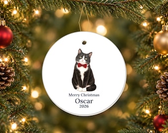 May include: A round white ceramic Christmas ornament featuring a black and white tuxedo cat wearing a red bow tie. Below the cat, the text reads Merry Christmas, followed by the name Oscar and the year 2026 in dark blue lettering.