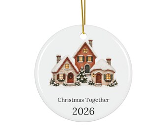 May include: A round white ceramic ornament featuring a painted illustration of three snow-covered houses with warm glowing windows and a central evergreen tree. The text reads Christmas Together 2026 in black font. A gold hanging cord is attached.