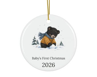 May include: A round white ceramic ornament featuring a black bear cub wearing a mustard yellow sweater and blue scarf, playing in the snow with pine trees. The text reads Baby's First Christmas and the year 2026 in black font.
