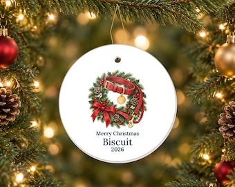May include: A round white ceramic Christmas ornament featuring a festive wreath illustration with a red dog collar and leash. The text reads Merry Christmas, Biscuit, and 2026 in dark blue lettering, centered below the wreath graphic.