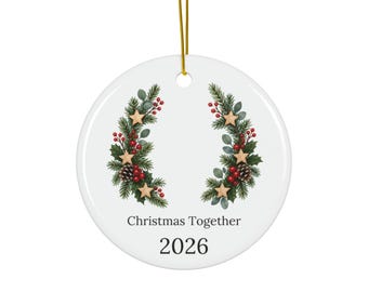 May include: A round white ceramic ornament hanging from a gold string. The design features two symmetrical evergreen branches adorned with red berries, pinecones, and gold stars. The text reads Christmas Together and the year 2026 in black font.