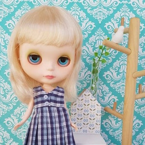 May include: A doll with blonde hair wearing a blue and white plaid dress and white shoes. The doll is standing on a wooden surface with a small cat figurine in front of it.