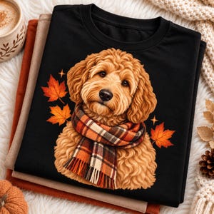 May include: A black crew neck t-shirt featuring a graphic of a golden-brown curly-haired dog wearing a plaid scarf. The dog is surrounded by orange and red autumn maple leaves and small four-pointed star accents on the front of the shirt.