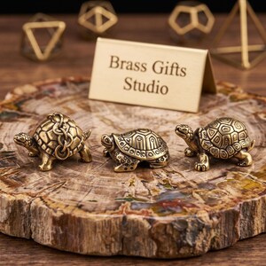 Puede incluir: Tres pequeñas figuras de tortugas de latón con patrones de caparazón detallados, una de ellas con una pequeña cadena adjunta, dispuestas frente a un letrero de metal dorado grabado con el texto Brass Gifts Studio, sobre una rodaja de madera petrificada.