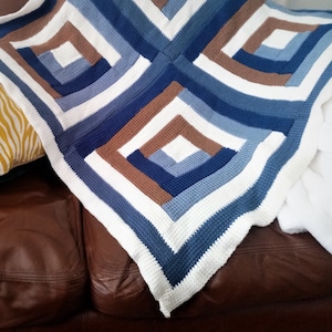 Può includere: Una coperta all'uncinetto bianca con quadrati blu e marroni. La coperta è piegata e drappeggiata su un divano in pelle marrone.